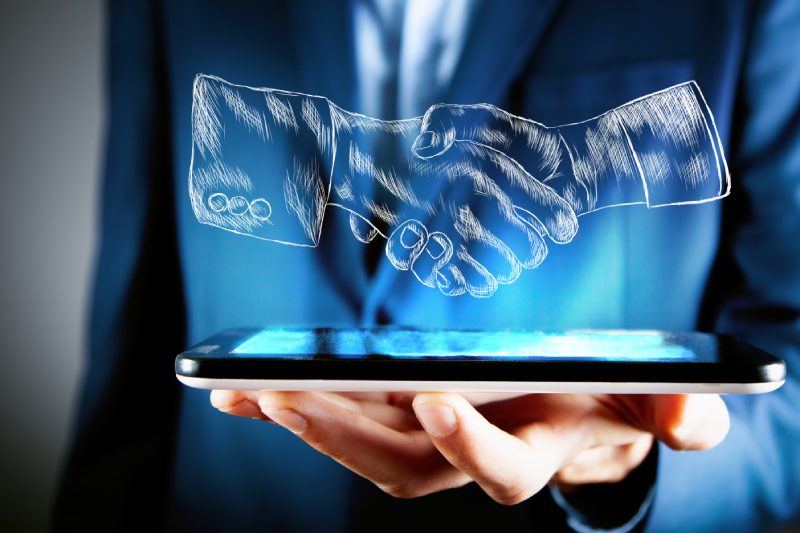 Deux mains holographiques semblant être en accord au-dessus d'une tablette tenue par un homme. L'image capture l'innovation et l'intégration technologique au cœur des opérations modernes, offrant un aperçu saisissant de la manière dont les réseaux hybrides transcendent les limites traditionnelles pour favoriser la connectivité et l'efficacité.