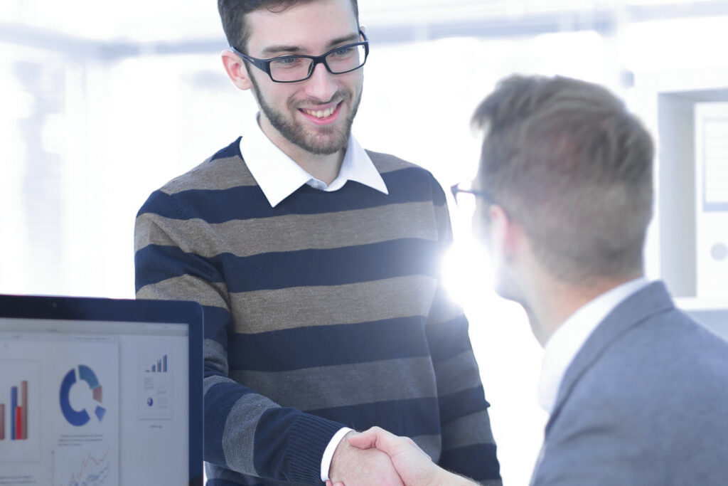 Un expert comptable souriant serre la main de son client, symbolisant une relation de confiance et de partenariat fructueuse. À leurs côtés, un ordinateur affiche des données chiffrées et des graphiques, soulignant l'utilisation des outils modernes dans un cabinet d’expert comptable ultra-moderne. L’image capture l'essence de la collaboration client-expert et l'intégration des technologies dans le domaine de la comptabilité.