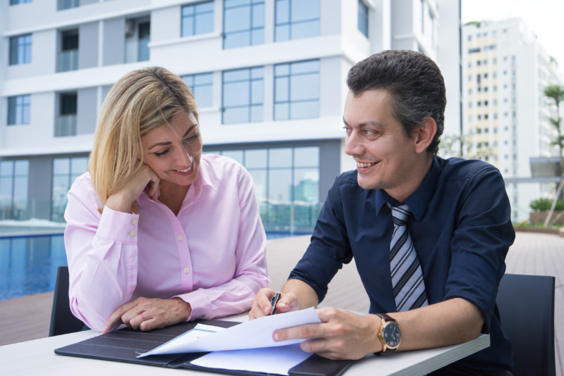 Une scène de négociation captivante montre un homme empathique expliquant et écoutant attentivement une femme à ses côtés. Le cadre extérieur comprend une terrasse avec en arrière-plan un immeuble moderne et vitré, évoquant une discussion professionnelle stratégique dans un environnement contemporain et ouvert.