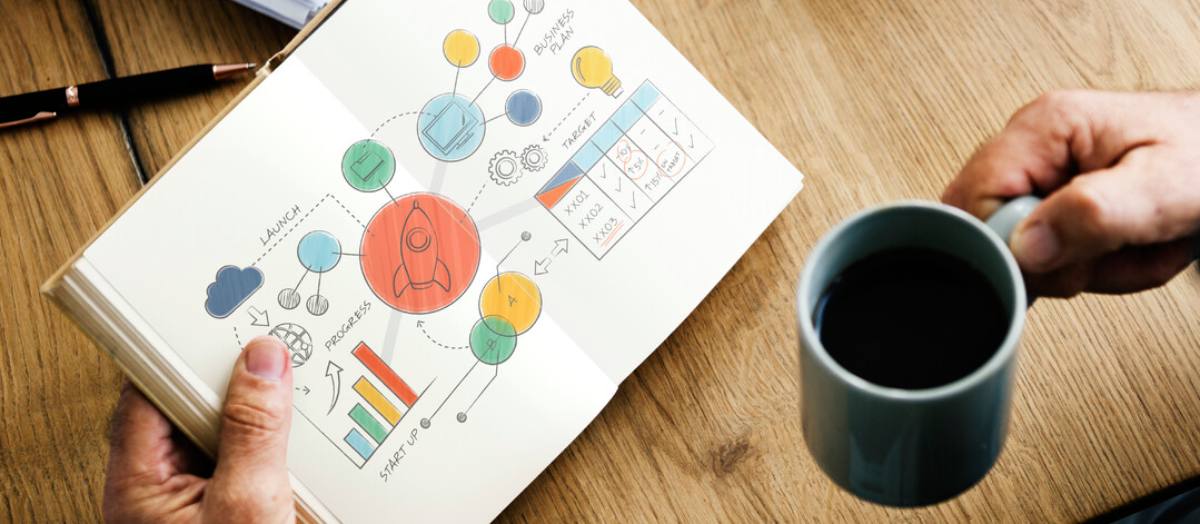 Une main gauche tenant fermement un agenda ouvert, révélant des pages remplies de schémas et de plans d'entreprise à long terme. Les diagrammes soigneusement dessinés et les notes détaillées indiquent une planification stratégique méticuleuse.